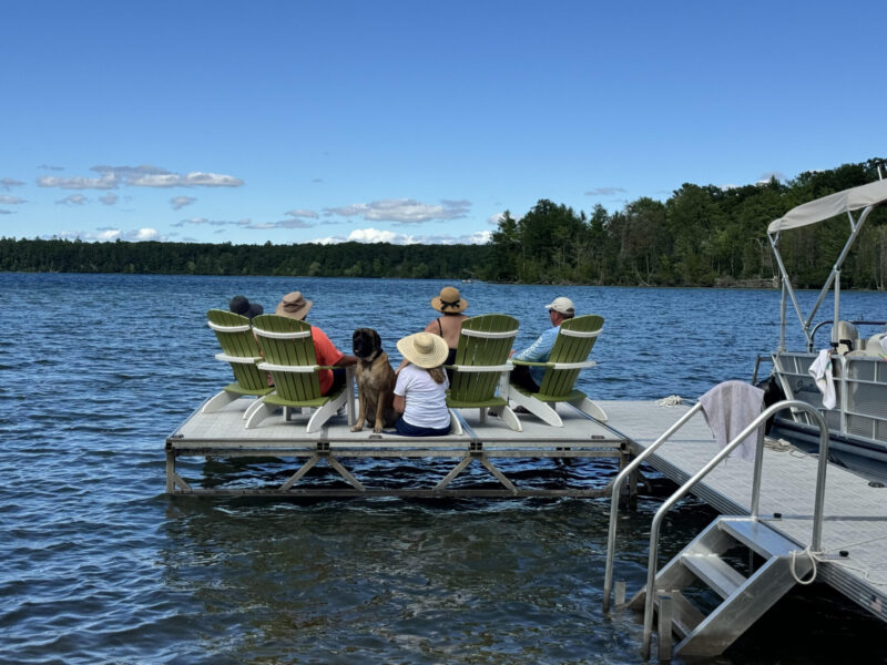 A photo of a lake shows a family sitting on a dock in green chairs with a dog. The photo is taken from behind the family, so it shows their backs as they look out onto the lake, which is under nearly cloudless blue skies.