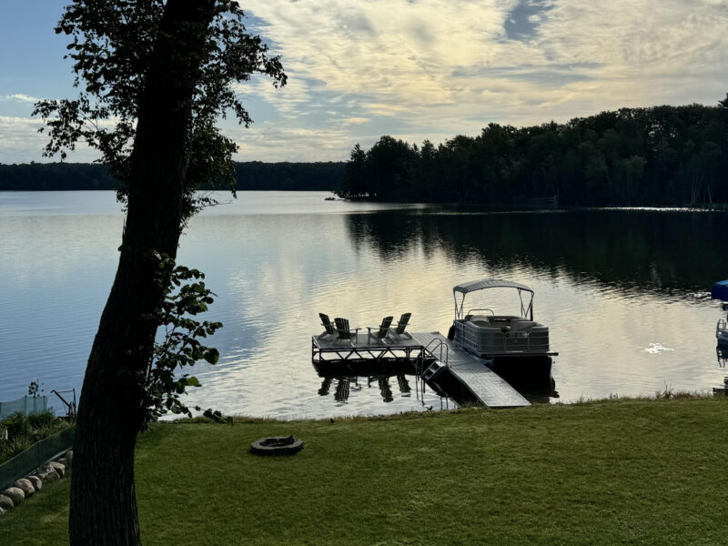 A serene morning setting on a lake, including a pontoon boat next to a dock, and a hazy blue sky that reflects off the water. 