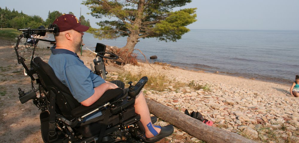 A man in a blue short-sleeve shirt and maroon cap sits in his wheelchair. He's seen from the side, at the edge of wooden rail fence and overlooking what appear to be seashells at the shore, with a body of water beyond on the right side of the frame. We see half of someone on the shore, and pines are in the background — including one on the shore.