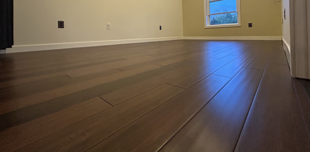 A picture of a bare, dark brown floor with vertical planks. A window is at one end of the room.