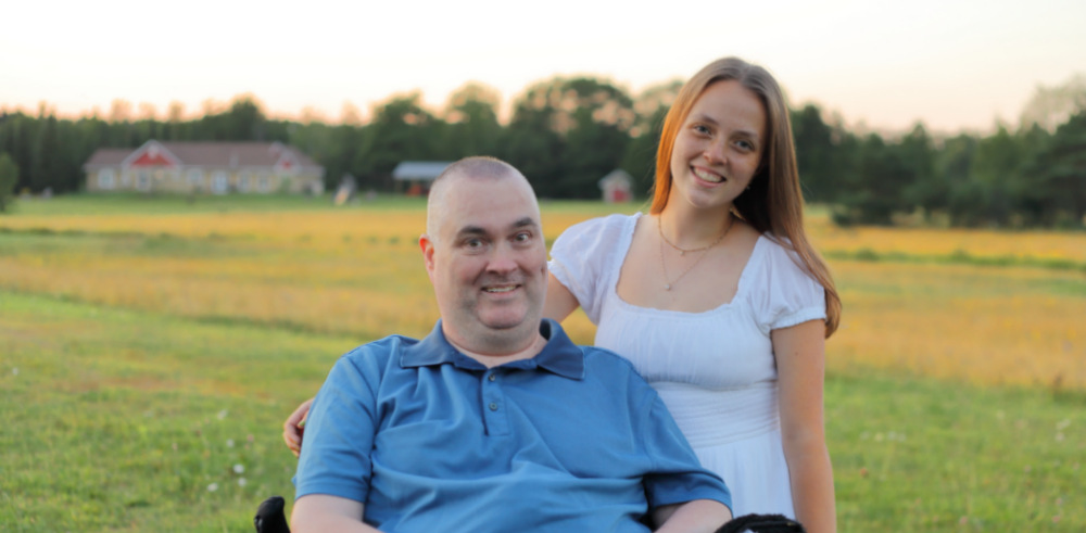 In a field of green and yellow, a man in a blue shirt sits in a wheelchair with a young woman - his daughter - in a white shirt standing beside him. She has her arm around the back of his shoulders.