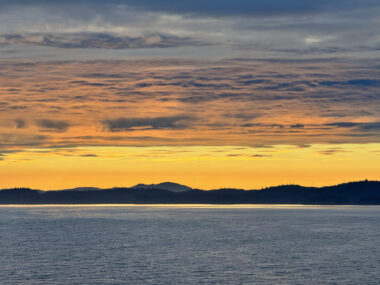 A photo taken from a cruise ship looks shows a sunset over the Alaskan coast. The water is a deep blue, and though the sun has gone below the horizon, the sky is golden and the clouds have taken on a beautiful orange hue.