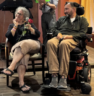 A man and woman are seated side by side on a wooden floor. The man is in a power wheelchair, while the woman is in a regular chair and holding a microphone and a red rose. Both are looking off to the right, as if at someone asking them a question.