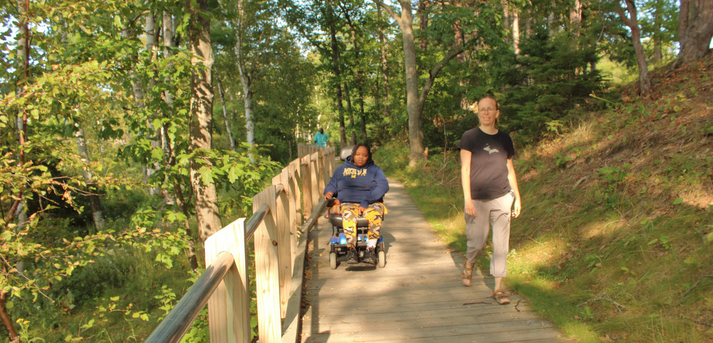 Two women navigate a boardwalk path through a wooded area. One is walking, while the other is in a power wheelchair.