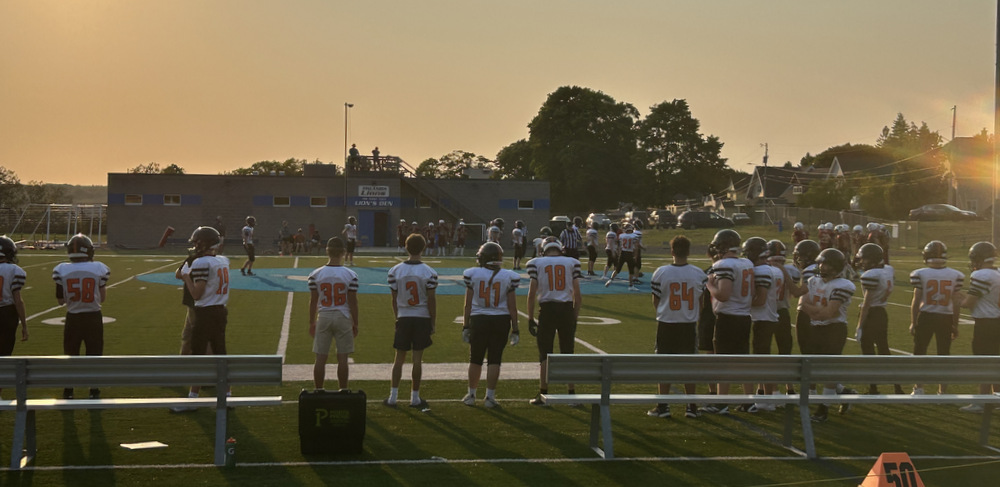 A photo taken from the bleachers shows a row of high school football players standing on the sideline of a field. Other players look to be lining up on the field, and the sun is setting behind the camera.