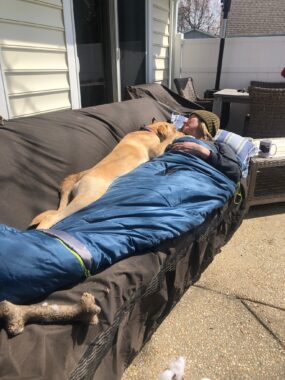 A woman lies on a couch on a patio outside her home. It's sunny outside, but it appears to be winter, as the patio furniture is covered and the woman is inside a blue sleeping bag and wearing a green knit beanie. Her head is turned to the side as she sleeps, and her dog is sprawled out next to her on the couch.