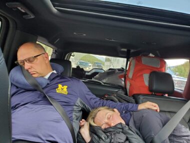 A photo taken from the front seat of a car shows a man and woman sleeping in the backseat. The man is sitting up but has his head propped on a small pillow atop his right shoulder. His left arm is resting on the woman, who is lying horizontally next to him with her head resting on what appears to be a rolled-up jacket. Luggage is in the space behind them.