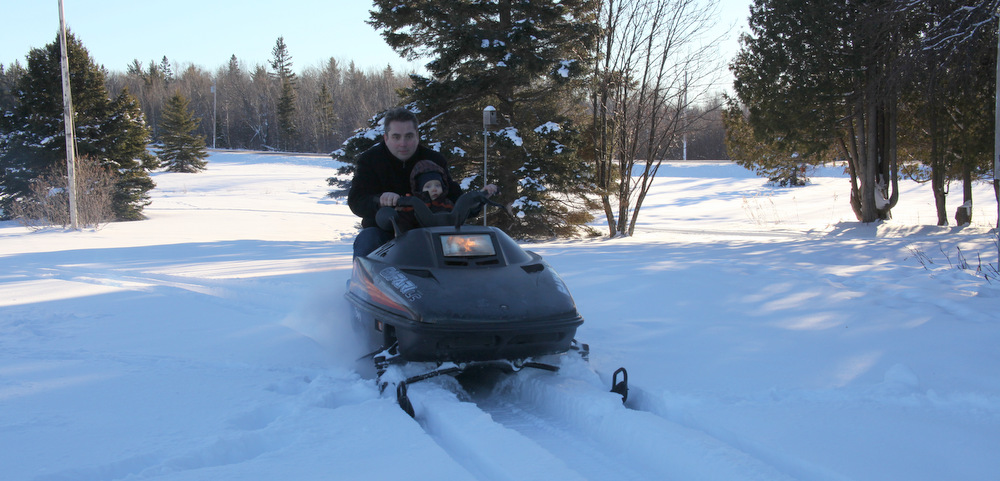 A 15-year-old photo shows an old, 1992 snowmobile being driven by a young man who has his 1-year-old son sitting in front of him. There is thick snow on the ground and pine trees scattered about in the background. 