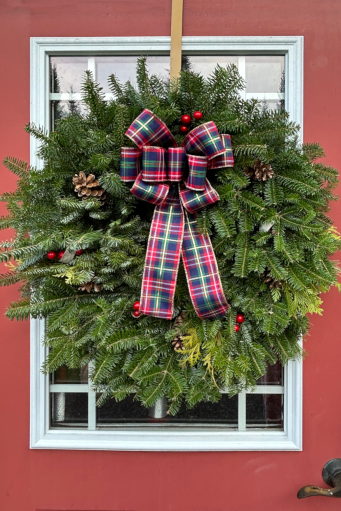 A beautiful handmade Christmas wreath hangs over a window on a red door. The wreath is made of boughs of Fraser firs and has a plaid ribbon in the center.