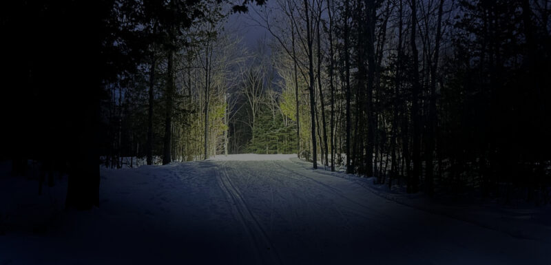 A dark ski trail bordered by tall trees is lit up in the distance.