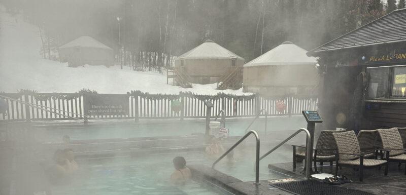 Several people soak in outdoor hot tubs at a Nordic spa in Michigan. There's a wooden structure on the right and several yurts in the background. It's wintertime, and the hill beyond the spa is covered in snow. Steam rises from the pools, suggesting it's extremely cold outside.