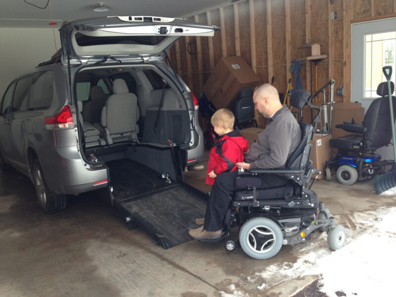 A man in a power wheelchair, with a young boy seated on his lap, navigates up a ramp into the back of an accessible van. 