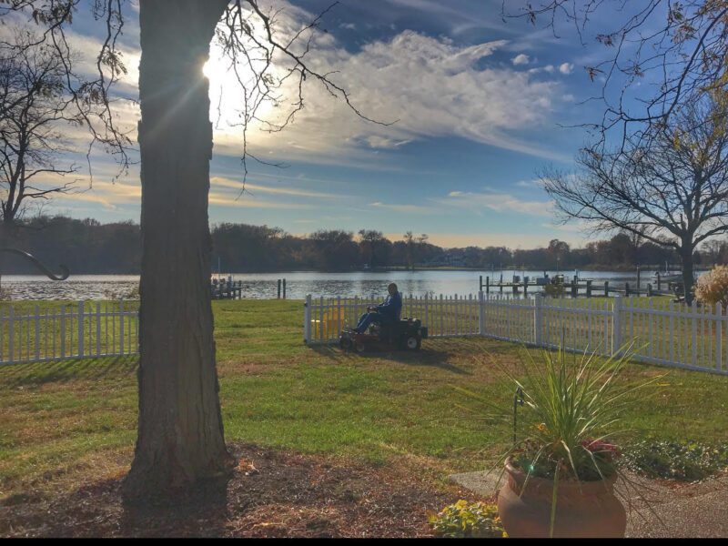 A scenic photo of a bright sun breaking through soft, white clouds in a blue sky. A man sits on a riding lawn mower and is mowing green grass. A lake is visible in the background, reflecting the blue of the sky. 