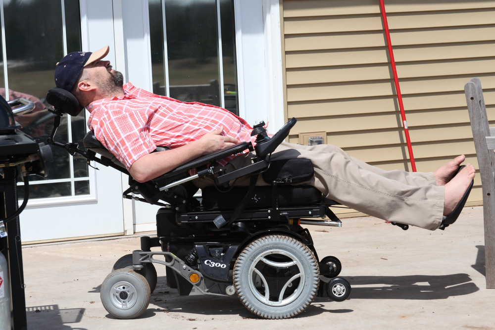 A man reclines in his power wheelchair, which is parked on the patio outside his home. He's wearing a light-red, button-down shirt and tan slacks, and has on a baseball cap to keep the sun out of his eyes.