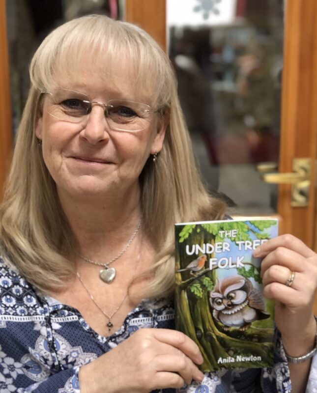 A woman probably in her 60s and wearing glasses holds up a book titled "The Under Tree Folk." The book has drawing of an owl standing on a tree branch on the cover. The woman is smiling.