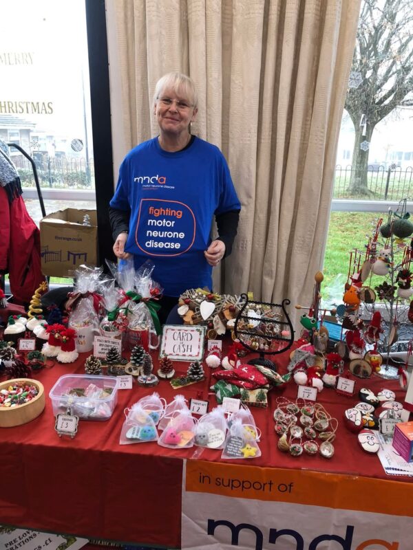 A woman demonstrates what's written on the front of her blue T-shirt. It says "Fighting Motor Neurone Disease." She stands behind a large table with a red table cloth covered in various Christmas-themed arts and crafts. 