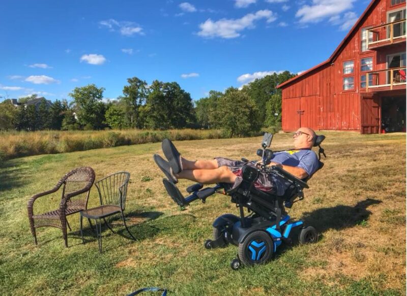 A vivid and brightly contrasted color photo of a rural, farm setting. A man is reclined in a power wheelchair located in broad field of cut grass, with a big red barn that looks like it's been converted into a home in the background. The sky is a deep, sunny blue, with sparse, white, fluffy clouds. Two chairs are sitting next to the man.