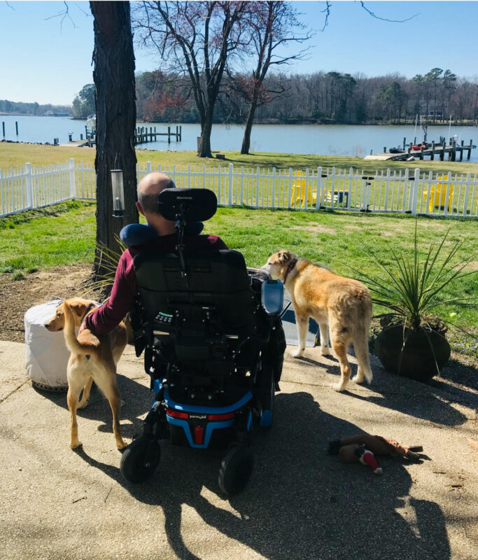A photo taken from behind a man in a power wheelchair as he looks out onto a vast yard that borders a large body of water, either a lake or a river. On the other side of this body of water is a forested tree line. On each side of the man are two golden-colored dogs. It's a sunny day, and the sun casts a long shadow behind the man's wheelchair.