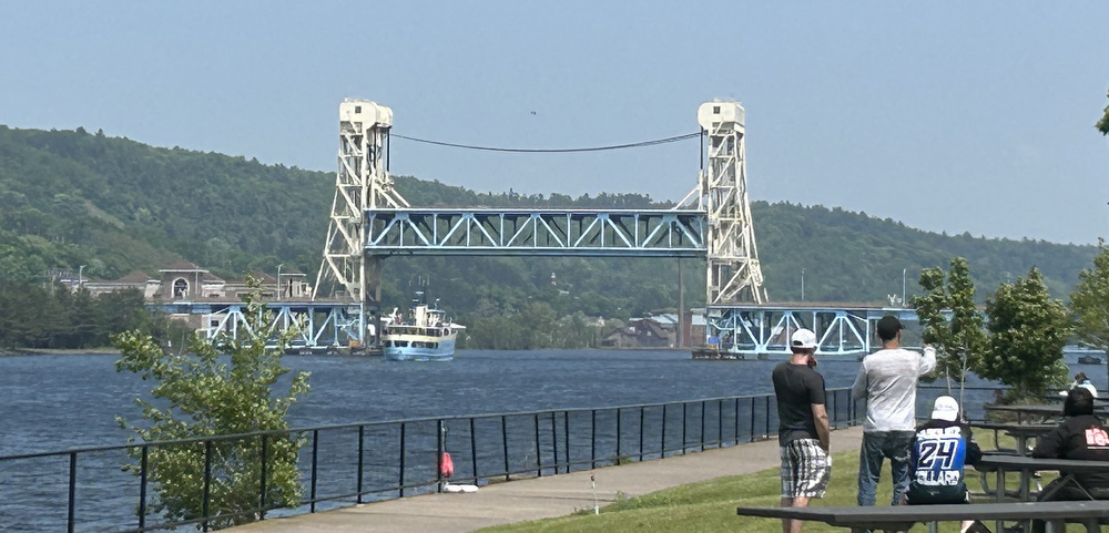 Under a blue sky with hills of greenery in the background, a white metal bridge stands in and over a waterway, its middle section raised for ships to pass beneath. To the right, on land and past a sidewalk, is an area of grass and picnic tables, with some people around.