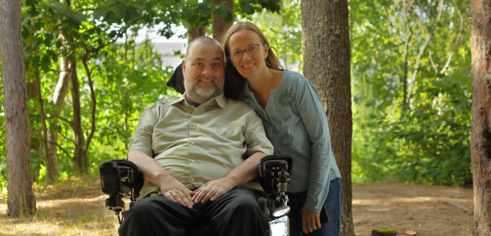 A couple in their late 40s smile for a photo outside, in the shade of several large trees. The husband is seated in a power wheelchair, and his wife stands beside him, leaning toward him with one arm around the chair.