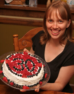 A smiling woman with light-brown, shoulder-length hair holds a glass plate holding a cake with white icing, decorated on top with an outside ring of blueberries and an inside of strawberries and raspberries, with blueberries again at center spelling out "30." 