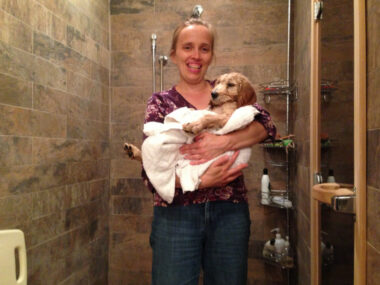 A woman stands in a nice shower stall of various shades of muted green and brown marbled tile. She holds a light-colored dog that's swaddled in a white towel.