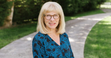 A blond woman with glasses poses for a photo on a shady sidewalk.