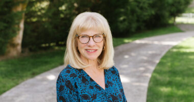 A blond woman with glasses poses for a photo on a shady sidewalk.