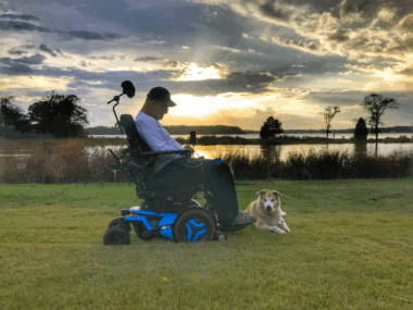 A man is pictured sitting in his power wheelchair on a lawn at sunset. He appears to be looking down at his feet, where a light-colored dog is lying. There's a large body of water in the background, but the man is sitting perpendicular to it.