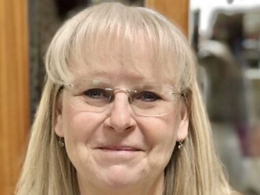 A close-up headshot of a blond woman with glasses.