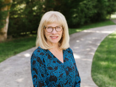 A woman stands on a shaded stone path with trees on one side and grass on the other. She is in the center of the frame and is smiling cordially, with a look (and glasses) to project a mix of intellectualism and humor.