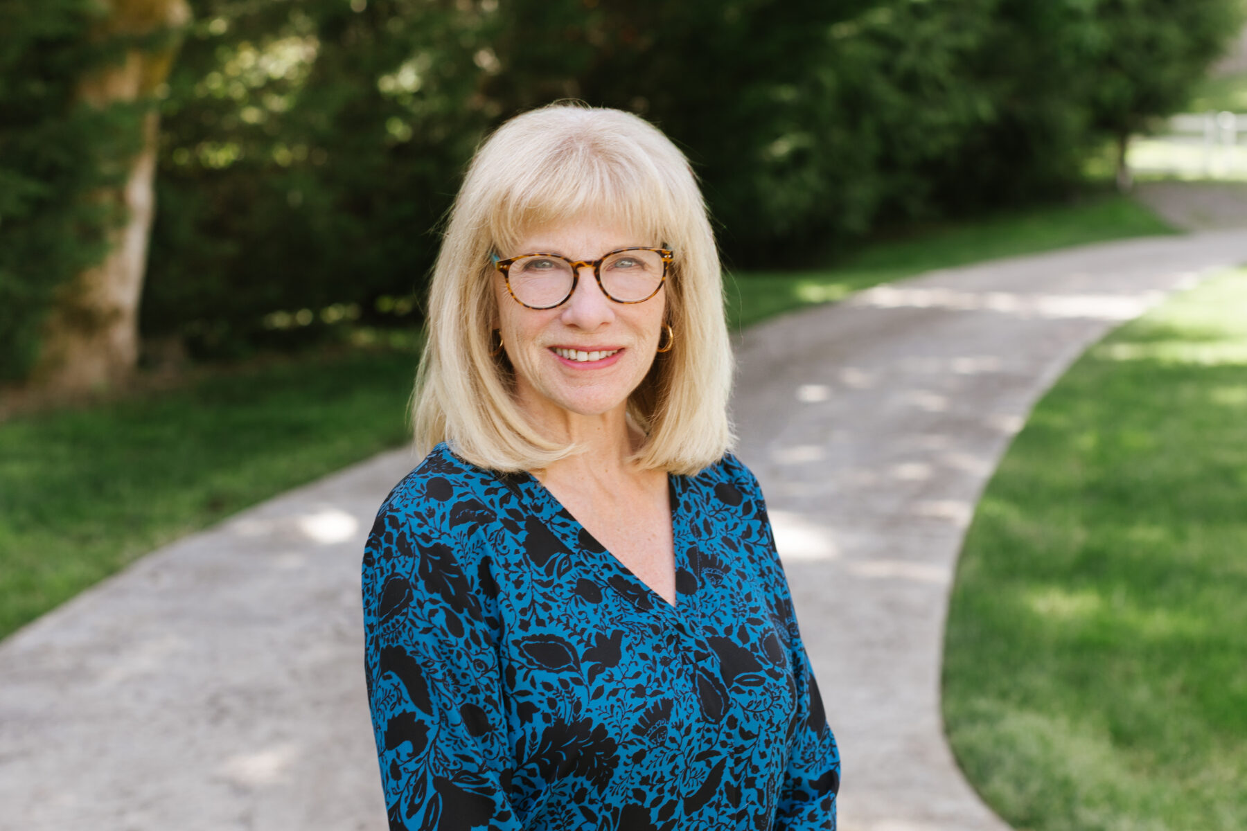 A woman stands on a shaded stone path with trees on one side and grass on the other. She is in the center of the frame and is smiling cordially, with a look (and glasses) to project a mix of intellectualism and humor.