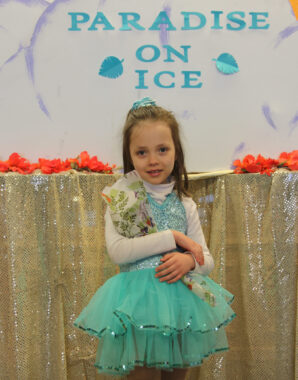 A young girl in an ice skating outfit clutches a bouquet as she stands in front of a sign that reads, "Paradise on Ice."
