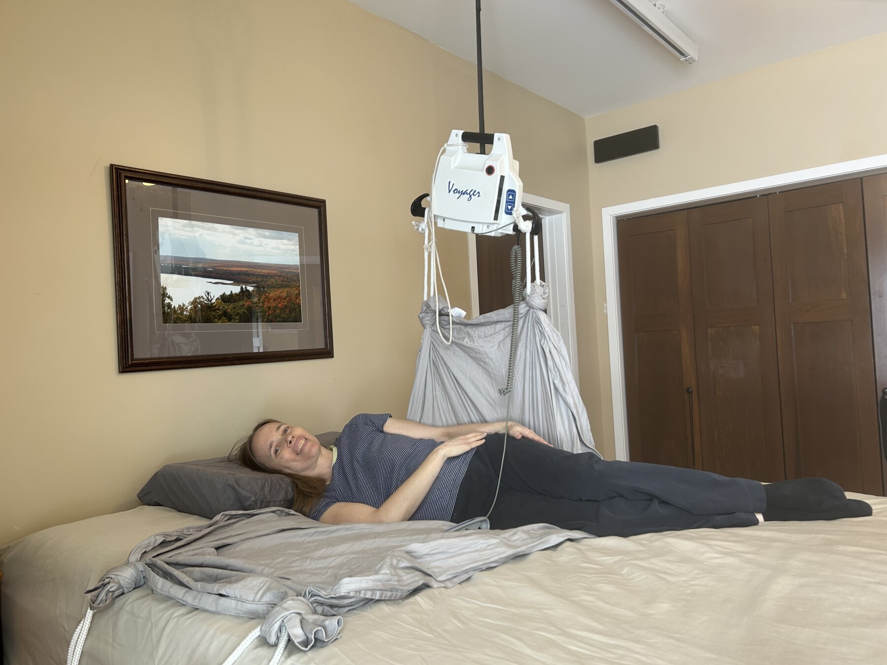 A smiling woman lies on a bed on top of a sheet with ropes affixed to it, and one end of the sheet is lifted into the air by an overhead lift machine. 