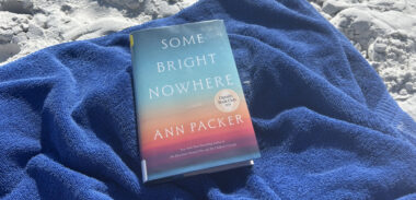 The novel "Some Bright Nowhere" sits on a blue beach towel on the sand.