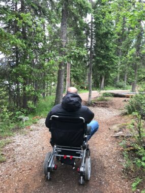 A photo taken from behind shows a man in a power wheelchair navigating a dirt path in a forest.