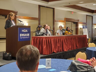 A woman stands at a podium with a "MDA Engage Symposium" plaque on the front. A group of people at a table are to the right, with one person speaking into a microphone. 