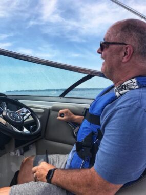 A man wearing a blue T-shirt, sunglasses, and life jacket sits in the driver's seat of a boat with his right hand on the throttle. He is smiling happily while looking out at the water.