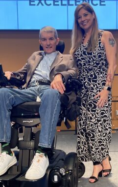 A man in a power wheelchair is next to a woman standing. It appears they're on a stage of some sorts in a large room. 
