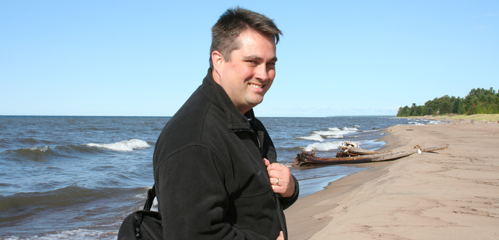 A man, pictured from the chest up, stands on the shore of a lake, smiling at the camera to his right. He's wearing a black coat and has a bag slung over his shoulder.